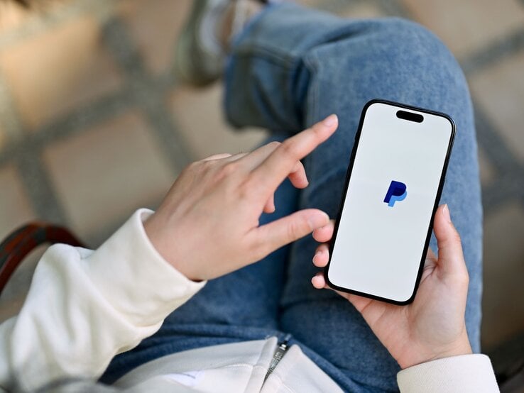 Eine Person in Jeans und weißem Pullover hält ein Smartphone mit dem PayPal-Logo auf dem Bildschirm und tippt mit einer Hand darauf.