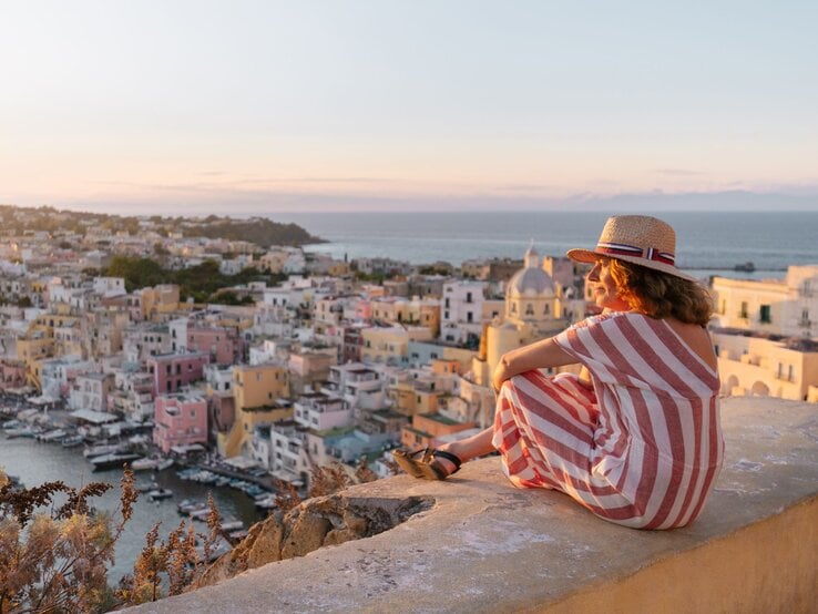 Zu sehen ist eine Frau mittleren Alters, die auf einer Mauer sitzt und auf eine mediterrane Stadt hinunterguckt.