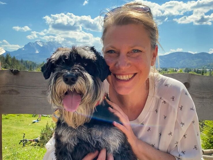 Fröhliche Frau, die einen kleinen, schwarz-weißen Hund im Arm hält. Sie befinden sich im Freien mit einer atemberaubenden Bergkulisse im Hintergrund. | © Oliver Nagel