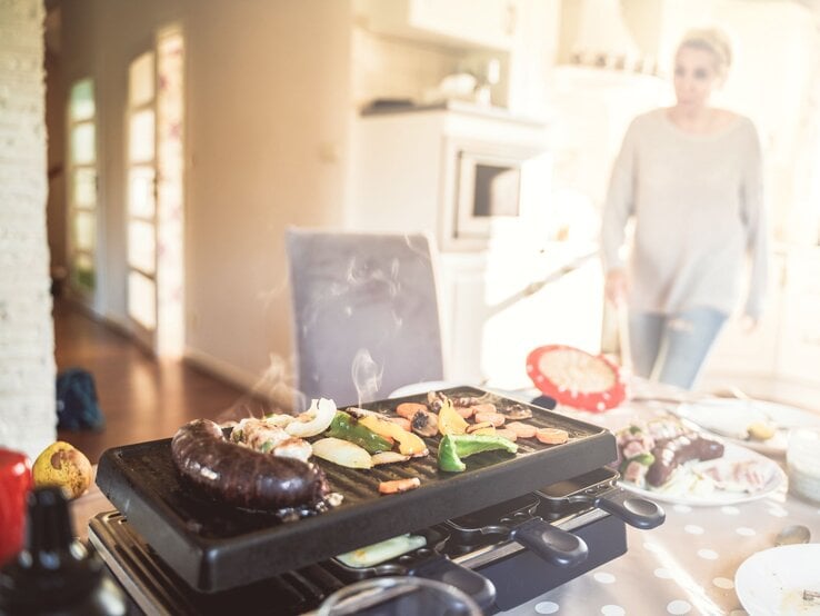 Ein Raclette mit rauchendem Gemüse und Würstchen steht auf einem gedeckten Tisch, im Hintergrund läuft eine Frau durch die Küche.