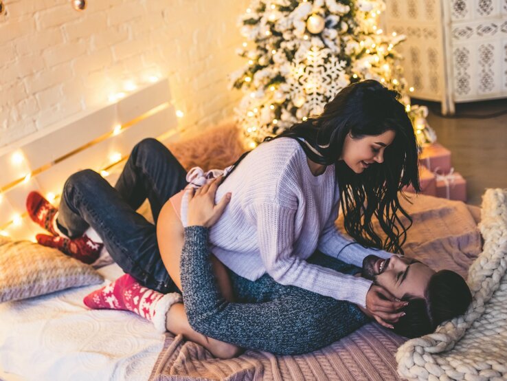 Ein Paar kuschelt verliebt auf einem Bett mit Lichterketten und Weihnachtsbaum im Hintergrund, beide wirken entspannt und fröhlich.