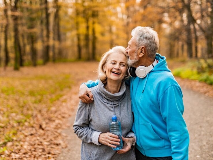 Ein älteres Paar spaziert lachend durch einen herbstlichen Wald, der Mann in blauer Sportkleidung küsst die Frau auf die Stirn.