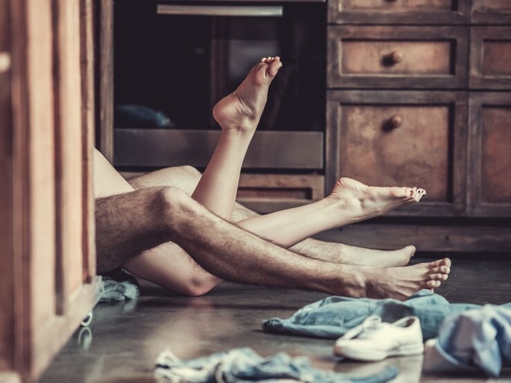 Zwei ineinander verschlungene Beine eines Paares liegen auf einem Küchenboden, umgeben von verstreuten Kleidungsstücken und Schuhen.