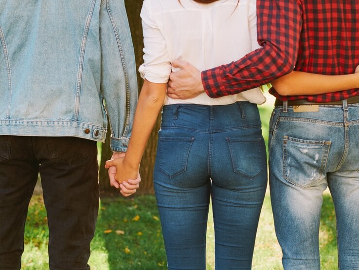 Drei Personen von hinten, die eng nebeneinander in einem Park stehen. Die mittlere Person, eine Frau, hält gleichzeitig die Hände von zwei anderen Personen an ihrer Seite.