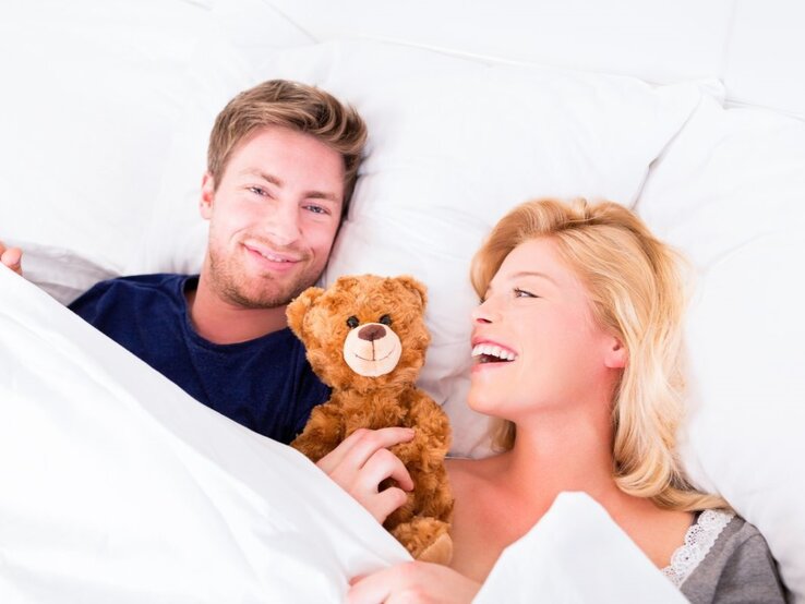 Paar im Bett, das sichtlich Freude hat. Der Mann hält einen braunen Teddybären und blickt in die Kamera. Die blonde Frau neben ihm lacht.