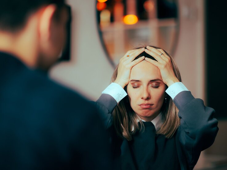 Frau und Mann in einer ernsten oder stressvollen Unterhaltung. Die Frau hält sich mit beiden Händen an den Kopf, die Augen geschlossen und einen besorgten Ausdruck im Gesicht. Der Mann, nur von hinten zu sehen, steht ihr gegenüber.