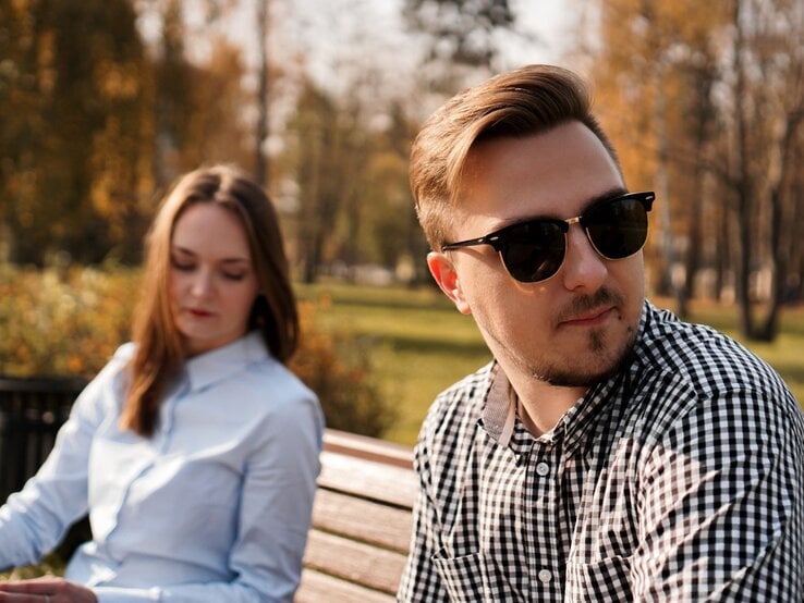 Junges Paar, das auf einer Parkbank im Freien sitzt. Der Mann trägt eine Sonnenbrille, schaut in eine andere Richtung und scheint gedankenverloren zu sein. Die Frau, die neben ihm sitzt, blickt nach unten und wirkt nachdenklich.