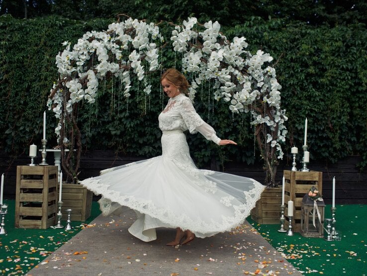 Eine lächelnde Braut mit hochgestecktem Haar dreht sich in einem weißen Spitzenkleid vor einem blumengeschmückten Hochzeitsbogen.