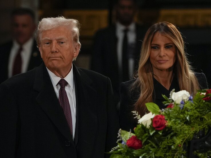 Ein älterer Mann mit grauen Haaren und schwarzem Mantel steht ernst neben einer Frau mit langen braunen Haaren und Blumenkranz in der Hand. | © IMAGO / Imagn Images