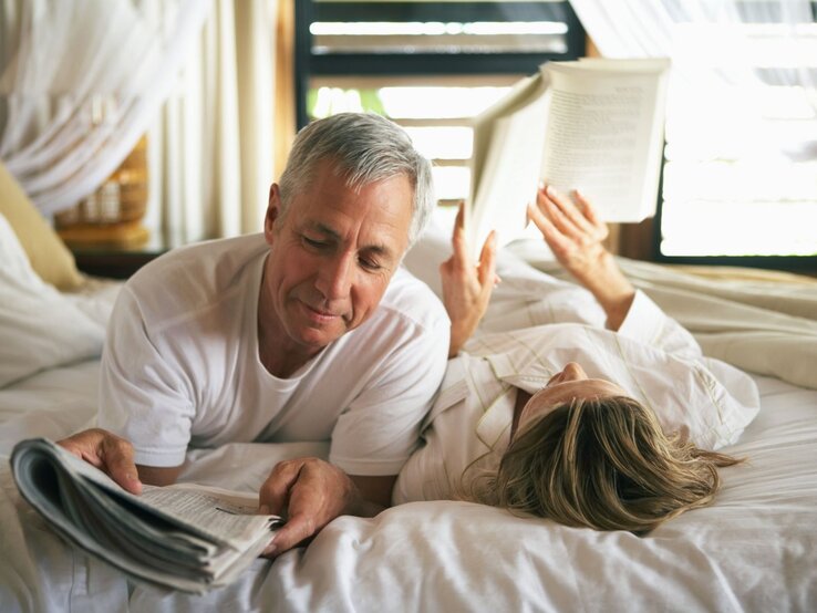 Reifes Paar liegt gemeinsam auf dem Bett, beide haben etwas zu lesen in den Händen, sie wirken glücklich und vertraut.