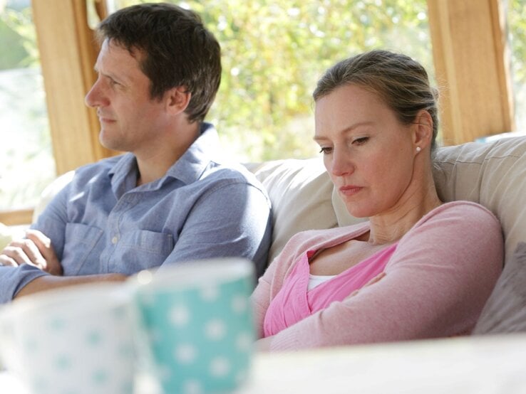 Ein Mann in blauem Hemd und eine Frau in rosa Oberteil sitzen mit verschränkten Armen schweigend auf einem Sofa und blicken angespannt weg.