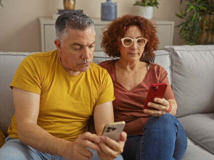 Ein mittelaltes Paar mit Smartphones sitzt konzentriert auf einem grauen Sofa. Der Mann trägt ein senfgelbes Shirt, die Frau eine Brille und rostrote Bluse.