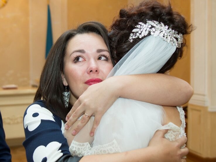 Eine Braut mit funkelndem Haarschmuck und Schleier umarmt eine weinende Frau in einem dunkelblauen Kleid mit weißen Mustern.