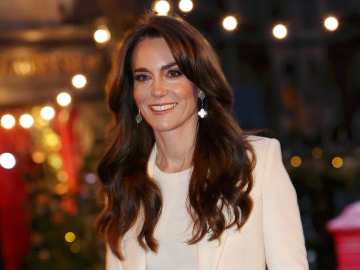 Prinzessin Kate mit langen braunen Haaren und Ohrringen lächelt vor einem festlich beleuchteten Hintergrund.