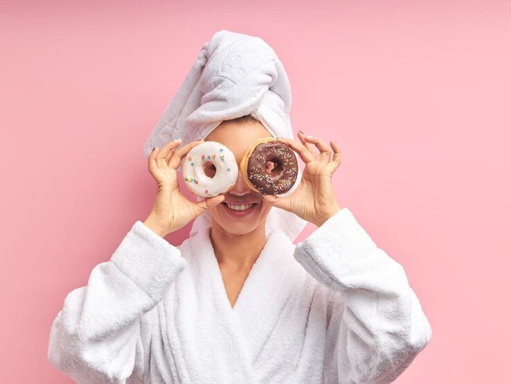 Frau in Bademantel und Handtuch lacht und hält Donuts mit Glasur und Streuseln vor ihren Augen vor rosa Hintergrund.