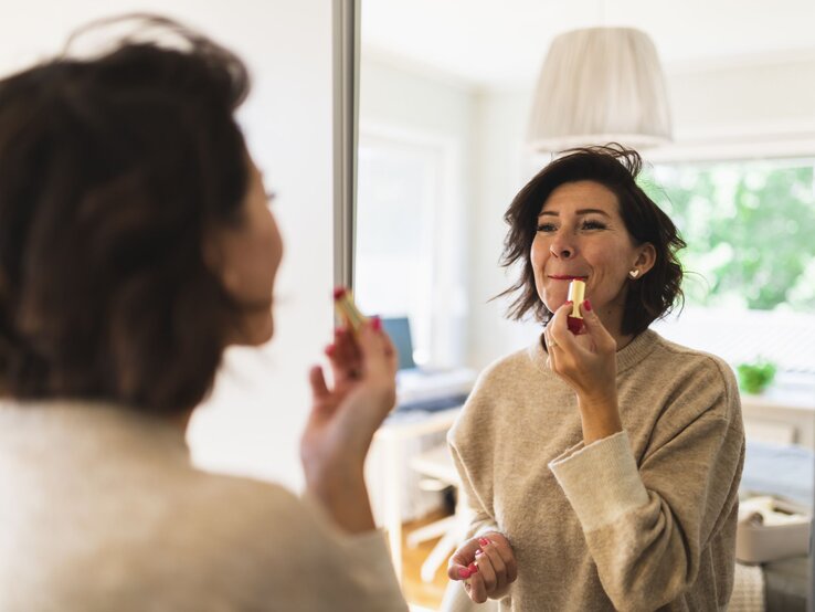 Frau im beigen Pullover trägt Lippenstift vor einem Spiegel auf, im Hintergrund ein gemütliches, gut beleuchtetes Wohnzimmer.