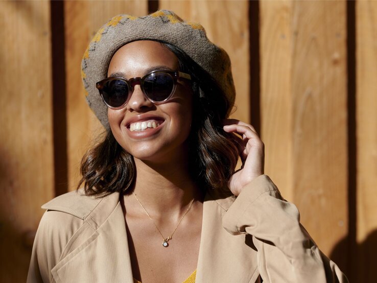 Lachende Frau mit Sonnenbrille und grauer Baskenmütze, in einem beigen Trenchcoat vor Holzwand im Sonnenlicht. | © Getty Images/ F.J. Jimenez