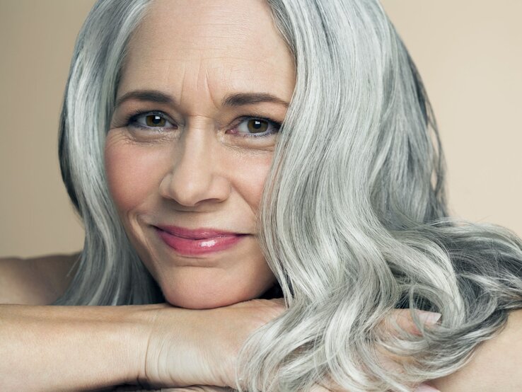 Reife Frau mit langen grauen Haaren und einem sanften Lächeln stützt ihr Kinn auf die Arme, Hintergrund beige. | © Getty Images/ Andreas Kuehn