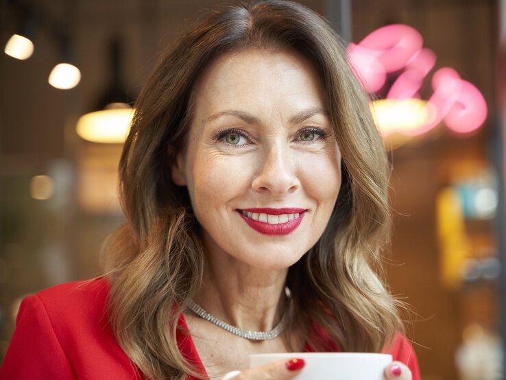 Lachende Frau mit rot lackierten Nägeln, rotem Lippenstift und Blazer, hält eine Tasse, sitzt in einem beleuchteten Café. | © Getty Images/ Westend61