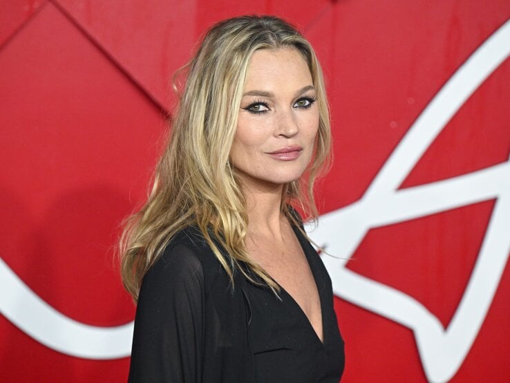 Kate Moss hat lange blonde Haaren, trägt dezentes Make-up und eine schwarze Bluse vor einem roten Hintergrund.