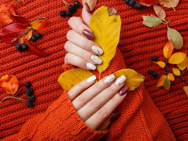 Hände mit herbstlich bemalten Nägeln halten gelbe Blätter auf einem orangefarbenen Strickpullover, umgeben von buntem Herbstlaub