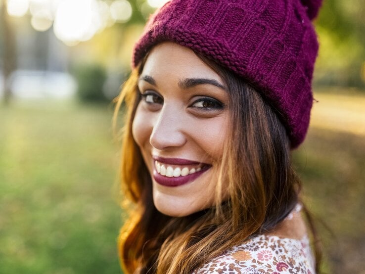 Junge Frau mit lebendigem Lächeln und bordeauxfarbener Strickmütze in herbstlicher Parklandschaft im goldenen Licht.