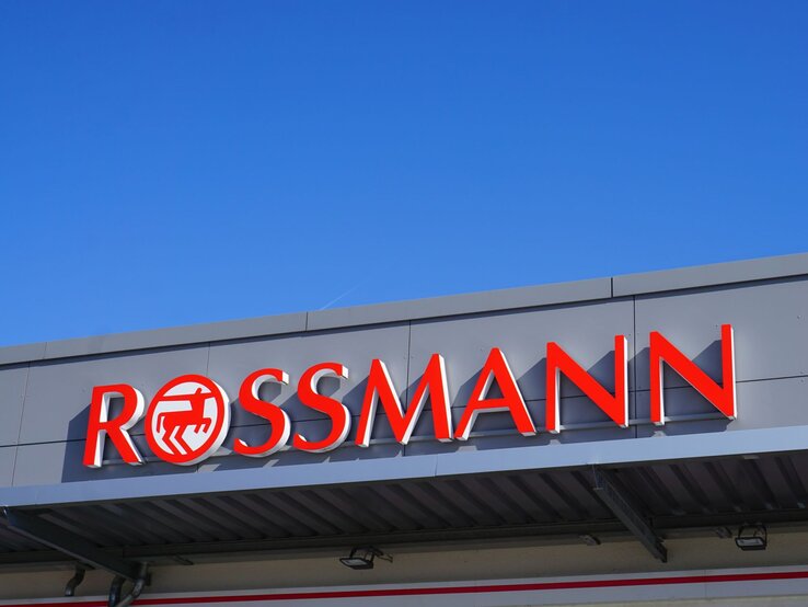 Rotes Rossmann-Logo auf grauer Fassade eines Geschäftes vor klarem, blauem Himmel.