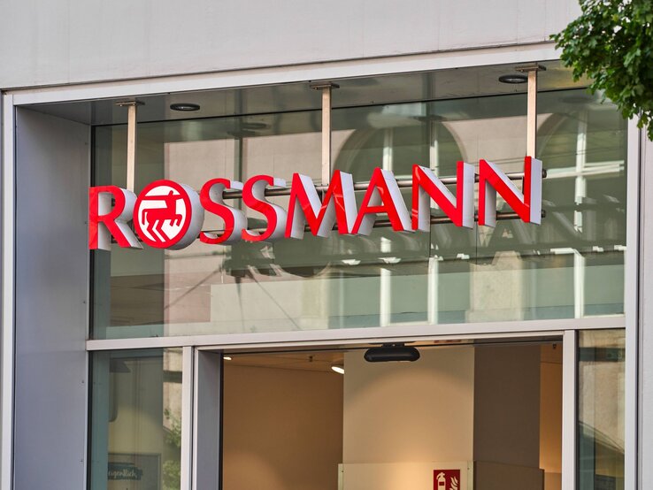 Außenansicht einer Rossmann-Filiale mit rotem Schriftzug und Glasfassade, dahinter unscharf erkennbares Innenleben.