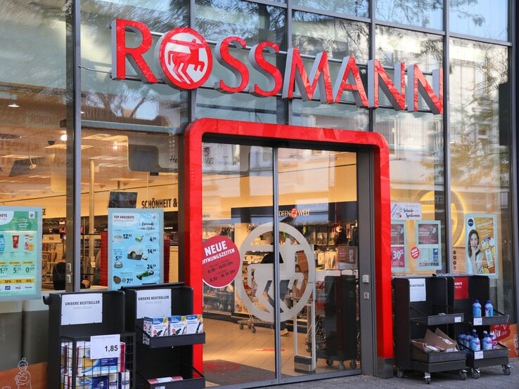 Frontansicht eines Drogeriemarktes von Rossmann mit Schaufensterplakaten, Einkaufswagen und Regalen mit Produkten vor der Tür.