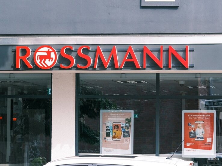 Eingang einer Rossmann-Filiale mit rotem Logo auf schwarzem Hintergrund, Schaufenster mit Werbeplakaten und einem parkenden Auto davor.