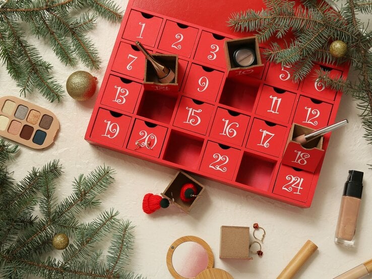 Ein roter Adventskalender mit Make-up-Artikeln, umgeben von Tannenzweigen, Lidschattenpalette und goldenen Kugeln.