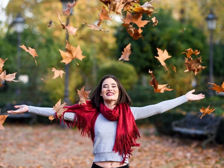 Eine junge Frau mit schulterlangem braunem Haar und rotem Lippenstift wirft lachend Herbstblätter in die Luft, umgeben von buntem Laub in einem Park.