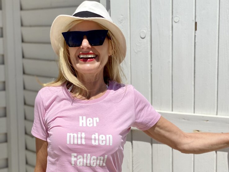 Lachende Frau mit Sonnenbrille und Hut, die ein rosa T-Shirt mit dem Aufdruck „Her mit den Falten!“ trägt. 