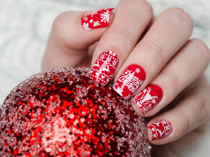 Hand mit roten, weihnachtlich verzierten Fingernägeln hält eine glitzernde rote Christbaumkugel vor einem hellen Hintergrund.