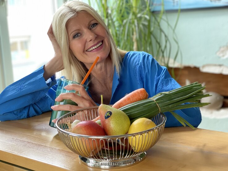 Fröhliche blonde Frau in blauem Oberteil trinkt einen grünen Smoothie mit Strohhalm, vor sich eine Schale mit Obst und Gemüse. | © Renate Zott