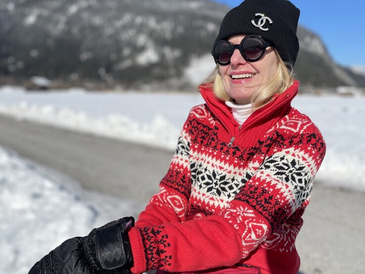 Lachende blonde Frau in rotem Norwegerpullover, schwarzer Mütze und Sonnenbrille sitzt mit Winterhandschuhen auf einer Bank im Schnee. | © Renate Zott