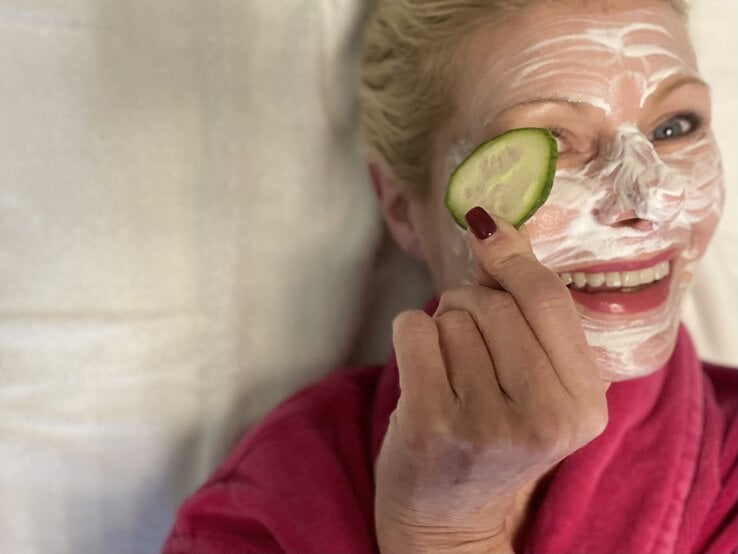 Eine lächelnde blonde Frau mittleren Alters in einem pinken Bademantel trägt eine Gesichtsmaske und hält eine Gurkenscheibe vor ihr Auge.