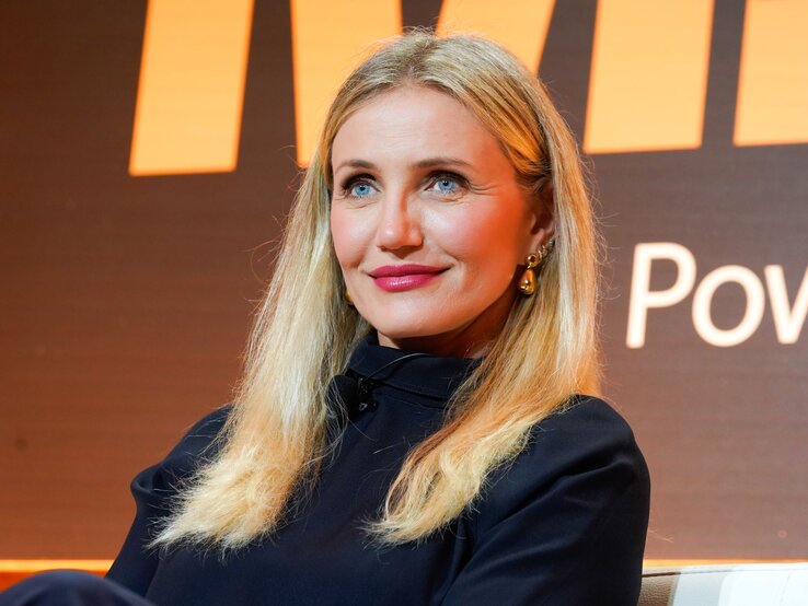 Cameron Diaz mit langen, offenen Haaren und goldenen Ohrringen lächelt dezent vor einem orange-schwarzen Hintergrund. | © Presley Ann/Getty Images for Fortune's Most Powerful Women Summit