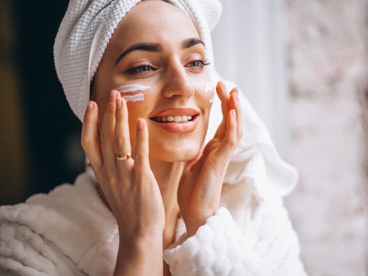 Nahaufnahme einer strahlenden Frau mit weißem Handtuchturban, die sich sanft Creme ins Gesicht einmassiert.