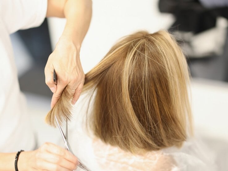 Person schneidet einer Frau die Haare. Das Haar ist blond mit dezenten Strähnchen und wird auf eine kurze Länge geschnitten, möglicherweise zu einem Bob.