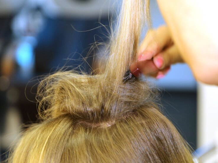 Eine Person toupiert den Haarwirbel auf dem Kopf einer Frau mit langem, blondem Haar, um Volumen zu erzeugen