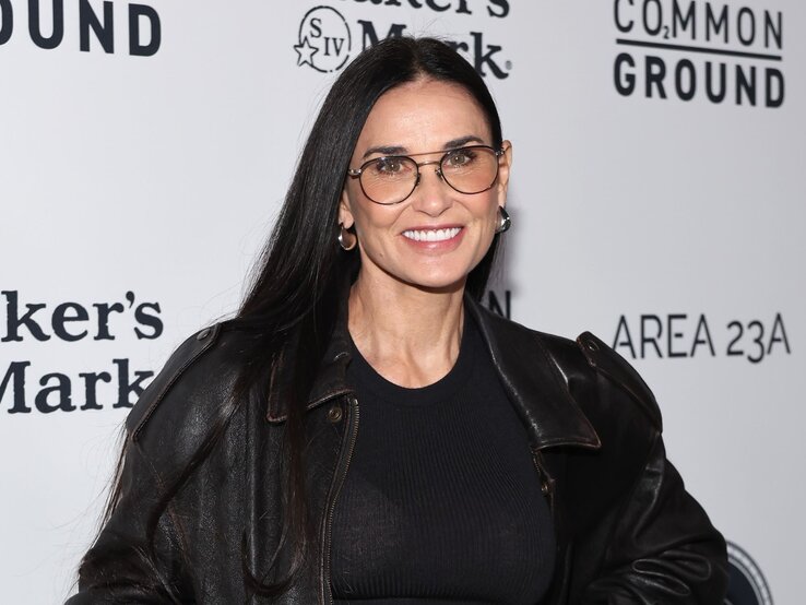 Demi Moore lächelt in die Kamera. Sie trägt eine schwarze Lederjacke und einen schwarzen Pullover, ergänzt durch eine große, runde Brille mit dünnem, braunem Rahmen. Ihr langes, dunkles Haar ist glatt und offen getragen. | © Getty Images / Amy Sussman