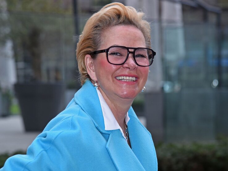 Claudia Effenberg lächelt in die Kamera. Sie trägt eine hellblaue Jacke und eine große, eckige Brille mit dickem, schwarzem Rahmen. Ihr blondes Haar ist zu einem eleganten, nach hinten gekämmten Stil frisiert. | © Getty Images / Tristar Media 