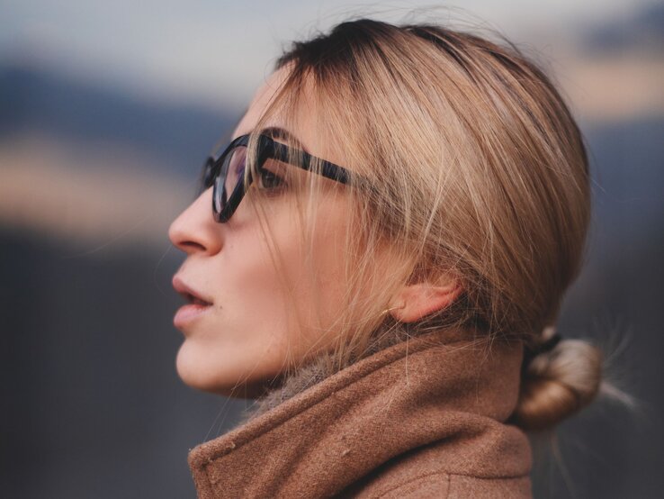 Junge Frau, die eine Sonnenbrille trägt und nachdenklich in die Ferne blickt. Sie hat blonde Haare, die zu einem lockeren Pferdeschwanz gebunden sind.