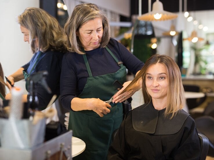 Eine Friseurin mittleren Alters schneidet einer lächelnden Frau mit glattem Haar die Spitzen im modernen Salon.