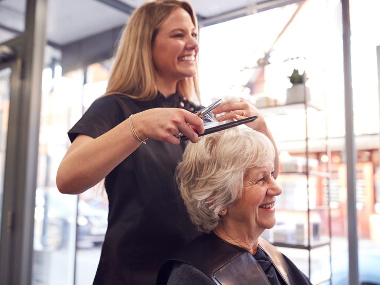 Eine junge Friseurin mit langen blonden Haaren schneidet lächelnd die Haare einer fröhlichen älteren Dame in einem hellen Salon.