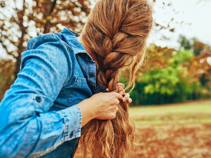 Zu sehen ist eine Frau in der Natur, die sich die Haare zu einem französischen Zopf flechtet