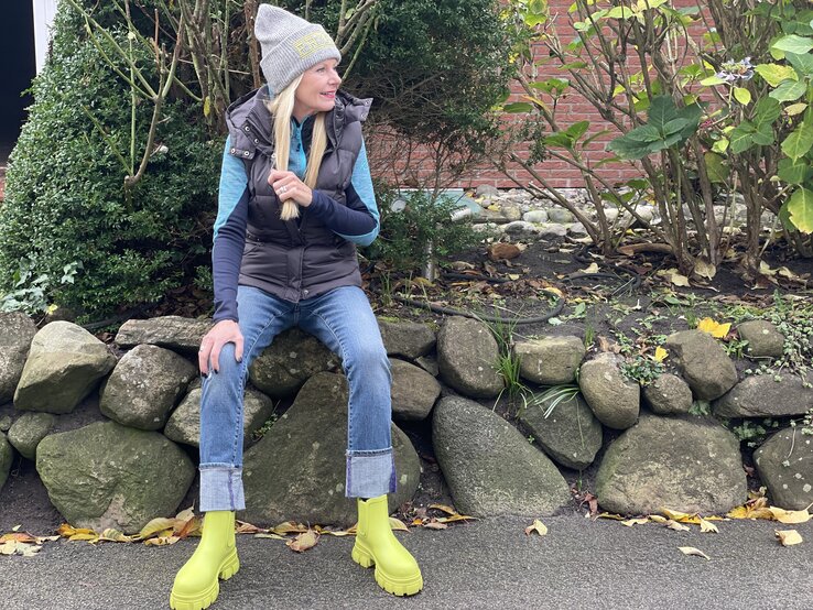 Frau, die auf einer Steinmauer sitzt. Sie trägt eine graue Mütze mit gelbem Schriftzug, eine gesteppte Weste, ein langärmliges blaues Shirt und hochgekrempelte Jeans. Ihre hellen, langen Haare fallen über die Schulter, sie hält eine Haarsträhne locker in der Hand. | © Renate Zott