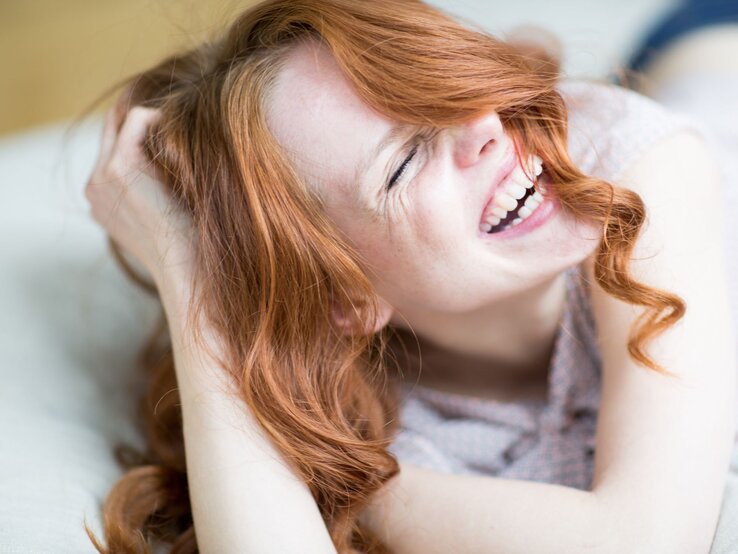 Junge Frau mit langen roten Locken lacht herzlich, während sie sich entspannt auf einem hellen Untergrund hinlegt.