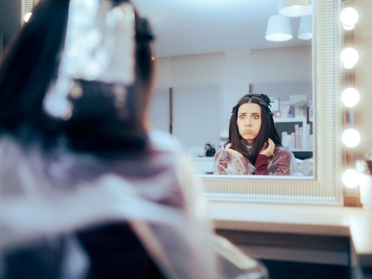 Frau im Frisiersalon, die besorgt oder erschrocken in den Spiegel schaut. Sie hat Frisiurzubehör im Haar und trägt einen Frisierumhang. Ihr Gesichtsausdruck deutet darauf hin, dass sie über das Ergebnis des Haarschnitts nicht glücklich ist. 